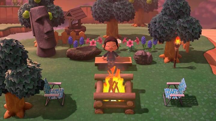 La joueuse est assise sur un banc de bois devant un feu de camp, entourée de décorations telles que des chaises pliantes, des flambeaux et une grande statue moaï.